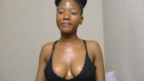 Media: Video of a young Black woman with medium brown skin and braided hair, wearing a black lace bra, standing against a plain beige wall, with a neutral expression.