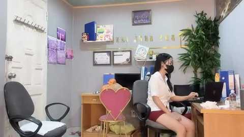 Media: Video of a modest office with a woman in a white blouse and mask seated at a desk, surrounded by a heart-shaped chair, computer, and potted plant.