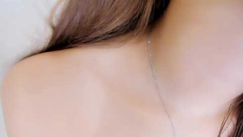 Video of a woman's bare shoulder and neck with light skin, long brown hair, and a delicate silver necklace.