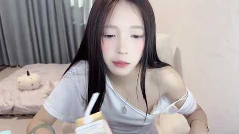 Media: A video of an East Asian woman with long black hair and pale skin, wearing a white off-shoulder top, applying makeup in a bedroom with a plush bed and gray curtains.