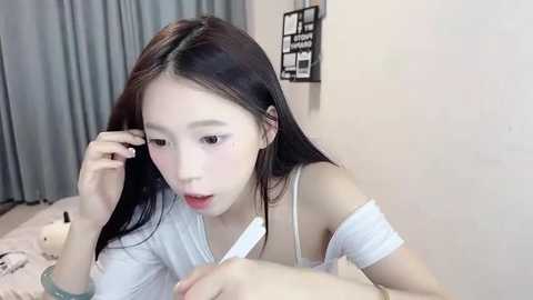 Media: Video of an Asian woman with long black hair, fair skin, and wearing a white off-shoulder top, looking surprised while holding her hair. Background features a grey curtain, a white wall, and a black-framed art piece.