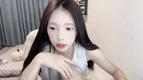 Media: Video of an East Asian woman with long, straight black hair, light skin, and fair complexion, wearing a white tank top, lying on a bed in a bedroom with beige walls and a gray curtain in the background.