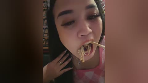 Media: Video of a young woman with medium brown skin, dark hair, and large eyes eating spaghetti with a fork, wearing a pink and white patterned shirt, in a dimly lit room.