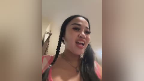 Media: Video of an Asian woman with long black hair in braids, wearing a pink and white striped top, smiling with her mouth open. Indoor setting with a beige wall and a lamp in the background.