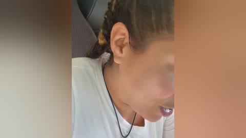 Media: Video of a young woman with medium brown skin, braided hair, wearing a white shirt, seated in a car, partially obscured by a person's arm.