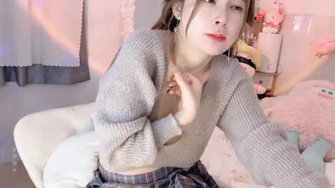 Media: Video of a young Asian woman with fair skin, wearing a grey knit sweater and a plaid skirt, sitting on a white sofa in a cozy, pink-themed room.