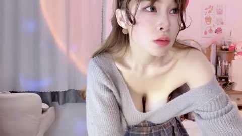 Media: Video of an Asian woman with light skin and long brown hair, wearing a loose, off-shoulder gray sweater, revealing ample cleavage. She gazes thoughtfully into the distance, set in a softly lit room with pastel decor.