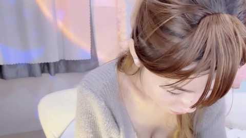 Video of a young woman with light brown hair styled in a bun, wearing a light gray sweater, sitting in a room with soft pastel curtains and a white table.