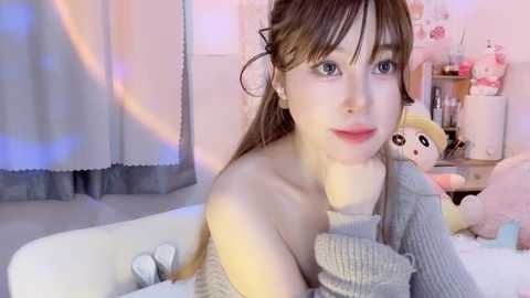 Media: Video of a young woman with fair skin, light brown hair, and blue eyes, wearing a grey off-shoulder sweater. She sits on a white couch in a softly lit, pastel-colored room with pink decor and stuffed toys.