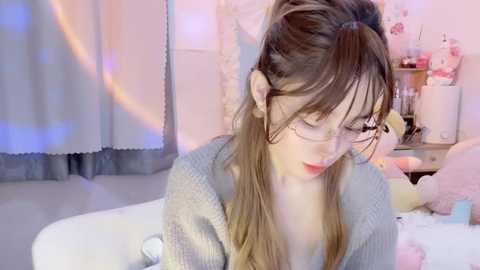 Media: Video of a young, fair-skinned woman with long brown hair, wearing glasses and a grey sweater, sitting on a bed with soft pastel decor.