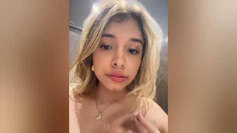 Video of a young woman with light brown skin, straight blonde hair, and large brown eyes. She wears a delicate necklace and a soft pink lip color. The background is blurred.