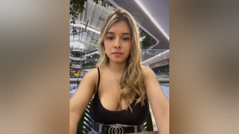 Media: Video of a young, light-skinned woman with long, wavy blonde hair, wearing a black spaghetti-strap top and a black belt, standing in a modern, indoor swimming pool area with glass railings and greenery.