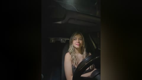 Media: Video of a blonde woman in a black dress, seated in a dimly lit car interior, looking directly at the camera with a neutral expression.