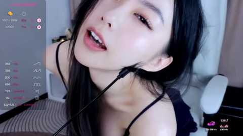 Media: Video of an East Asian woman with long black hair, wearing a black top, leaning forward with a headset microphone, in a modern, brightly-lit room with white furniture and a striped carpet.