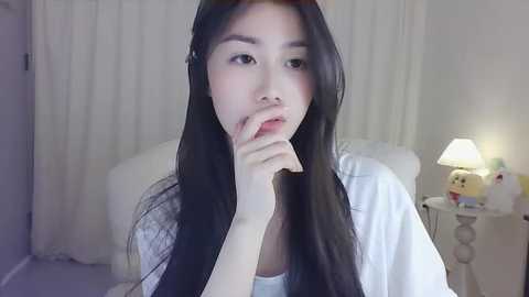 Media: Video of an Asian woman with long black hair, fair skin, and light makeup, holding her finger to her lips, in a softly lit bedroom with white curtains, a lamp, and a plush toy.