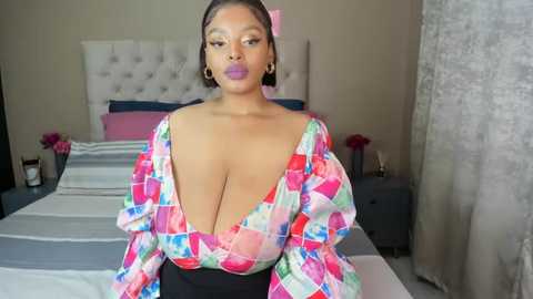 Media: Video of a curvy, dark-skinned woman with medium-length hair, wearing a colorful, low-cut, patchwork blouse, posing in a modern, beige-tiled bedroom with a tufted headboard.