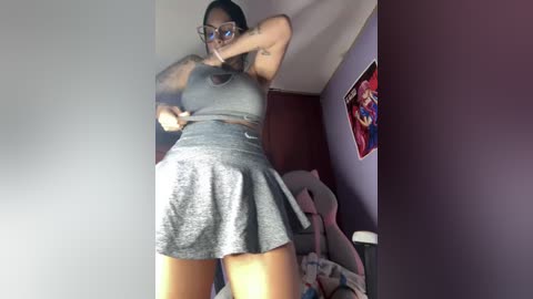 Video of a young woman with medium brown skin, wearing glasses, a gray sports bra, and a matching skirt, posing confidently in a dimly lit room with posters and a plush toy.