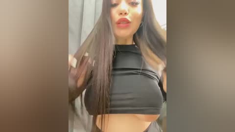 A video of a young woman with long, straight black hair, wearing a black crop top and red lipstick, posing in front of a blurred, beige background.