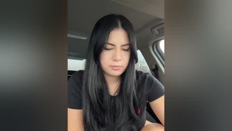 Media: Video of a young Latina woman with long, straight black hair, wearing a black T-shirt, seated inside a car. She has a neutral expression, looking down. The car's interior is dimly lit.