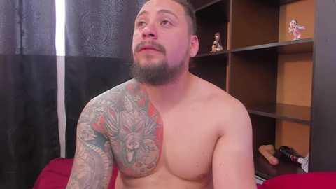 Video of a shirtless, muscular man with a beard and tattoos, including a large, colorful sleeve, sitting on a red couch. Background includes a black curtain, wooden shelves, and figurines.