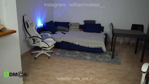 Media: Video of a small, cozy room with a white gaming chair, blue sofa, and a dark wooden desk. The floor is tiled in beige, and the walls are white.