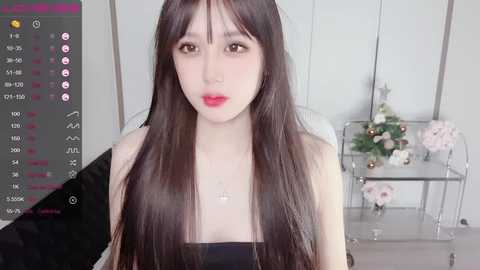 Media: Video of a young East Asian woman with long, straight black hair, fair skin, and a slender physique, wearing a black strapless top. Background features a glass shelf with flowers, a TV screen displaying a live stream interface.