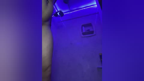 Media: Video of a nude woman with fair skin, medium build, and small breasts, standing in a dimly lit room with purple LED lights, under a showerhead.