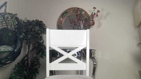 Media: A video showing a white wicker chair with a decorative floral arrangement on top, placed against a beige wall with a round mirror and green leafy plants.