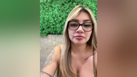 Media: Video of a young Caucasian woman with long blonde hair, wearing black-framed glasses, and a brown top, standing outdoors with a green hedge and gravel background.