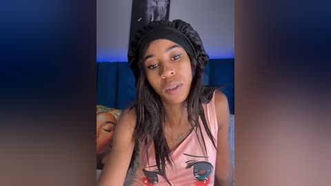 Media: Video of a young Black woman with long braids, wearing a black headscarf, a pink tank top with cat faces, and a serene expression. Background features a dark headboard and a blurred decorative pillow.