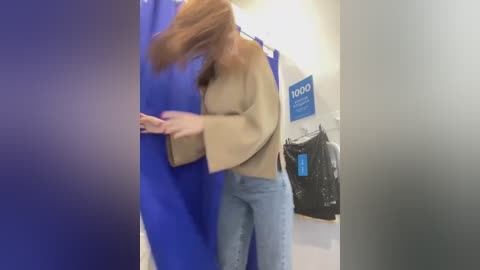 Media: Video of a woman in a beige sweater, blue jeans, and brown boots, walking quickly through a store, hair flying, with a blue curtain, a \"100%\" sign, and a black jacket hanging on a rack in the background.