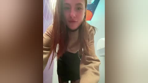 Media: Video of a young woman with long, straight, reddish-brown hair, wearing a beige sweater over a black top, leaning over a sink in a bathroom with a white and blue tiled wall.