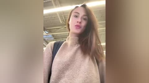 Media: Video of a young woman with light skin and long, straight auburn hair, wearing a beige turtleneck sweater and black backpack, taken indoors with exposed industrial ceiling.