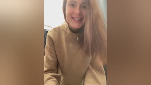 Media: Video of a young woman with long, straight blonde hair, wearing a beige sweater, smiling broadly, slightly out of focus, against a blurred brown background.