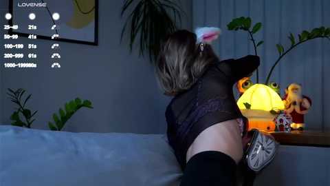 Media: Video of a woman in black lingerie and bunny ears, bending over a couch, in a dimly lit room with a glowing lamp and stuffed animals.