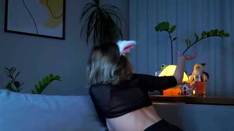 Media: Video of a woman with short blonde hair, wearing a black crop top and white bunny ears, sitting on a couch, playing a video game in a dimly lit room with potted plants and a lamp.