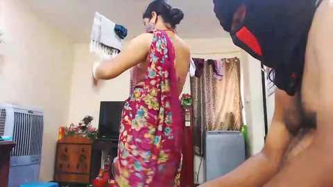 Media: Video of a woman in a red floral dress with a slit, holding a fan in a cluttered, dimly-lit room with a refrigerator, TV, and air conditioner.