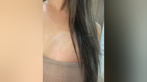 Video of a woman with long, straight, dark brown hair, wearing a beige, off-shoulder, ribbed top, partially cropped, with a blurred background.