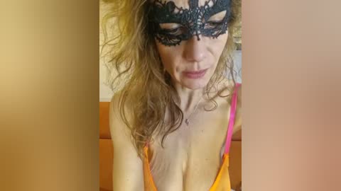 Media: Video of a woman with wavy blonde hair, wearing a black lace mask and a low-cut, orange dress, sitting on a light-colored couch.