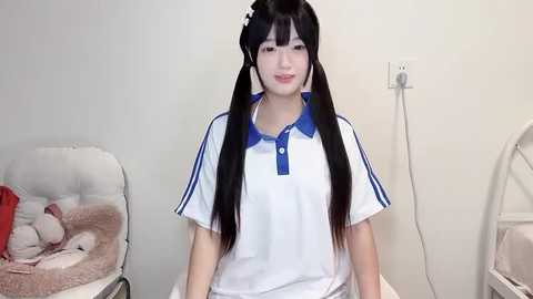 Media: Video of an East Asian woman with long black hair in pigtails, wearing a white polo shirt with blue trim, smiling. Background shows a beige chair with a red blanket, and a white electrical outlet.