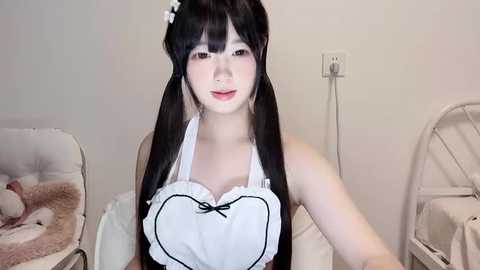 Media: Video of an East Asian woman with pale skin, wearing a white maid outfit with a black heart-shaped top, sitting in a beige room with a baby chair and a white fan.