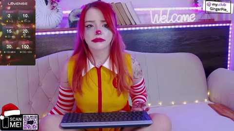 Media: Video of a fair-skinned, red-haired woman in a yellow dress with red stripes, sitting at a white couch with a keyboard. Background features a dark wooden wall with books and a \"Welcome\" sign.