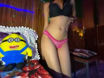 Media: Video of a slender woman with light skin wearing black lingerie and pink panties, standing in a dimly lit room with a Minion plush toy and a blue bedspread.