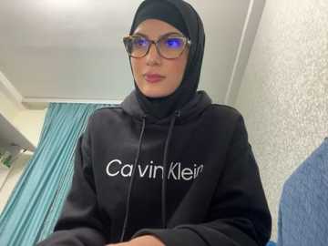 Media: Video of a woman in a black Calvin Klein hoodie with a hood, wearing glasses and a headscarf, standing indoors with teal curtains and a blue chair in the background.