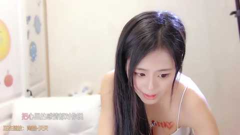 Media: Video of an East Asian woman with long black hair, wearing a white tank top, in a brightly lit bathroom with white tiles and colorful stickers.