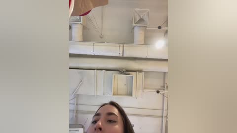 Media: Video of a young Asian woman with medium-length dark hair, partially obscured by white walls and air ducts in an industrial setting, with a red ceiling fan visible.