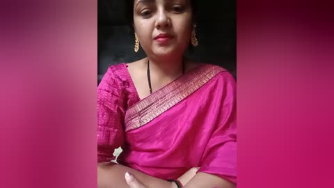 Media: Video of a South Asian woman with medium brown skin, wearing a bright pink saree with intricate embroidery and gold earrings, against a gradient pink background. She has a neutral expression, crossed arms, and her dark hair is tied back.