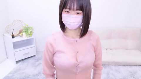 Media: Video of an East Asian woman with short, straight black hair, wearing a pink mask and long-sleeved top, showcasing large breasts. Background features a white couch, white shelves, and a plant.