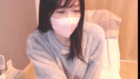 Media: Video of an Asian woman with long black hair, wearing a white face mask, grey sweater, and seated indoors with light wooden furniture and beige walls.