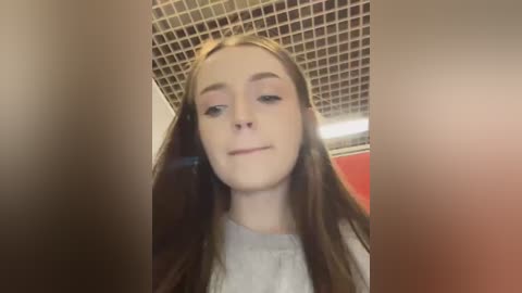 Media: Video of a young woman with long, straight brown hair, wearing a white shirt, standing in a bathroom with a tiled ceiling and a red wall in the background.